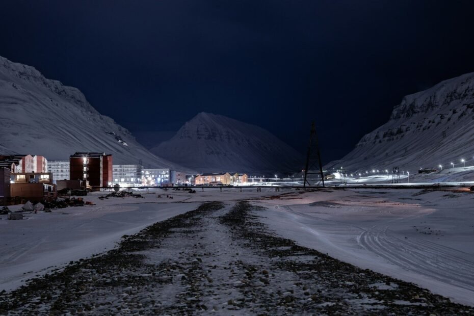 a snowy road at night with buildings and mountains in the background